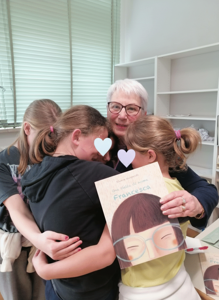 Immagine che raffigura una cliente di Radicilab sorridente, con il suo libro in mano e tre bambine che la abbracciano. Il volto delle bambine è nascosto da delle emoticon per motivi di privacy.